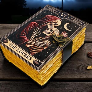 Op de afbeelding: Een tarotkaartjournaal met een zwarte kaft met een skeletpaar dat elkaar omhelst onder een halve maan en sterren. De kaart is gelabeld "THE LOVERS" en heeft een messing sluiting en verouderde, vergeelde pagina's. Het journaal ligt op een donker houten oppervlak.