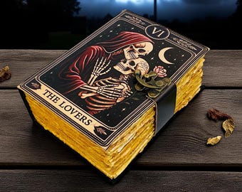 Op de afbeelding: Een tarotkaartjournaal met een zwarte kaft met een skeletpaar dat elkaar omhelst onder een halve maan en sterren. De kaart is gelabeld "THE LOVERS" en heeft een messing sluiting en verouderde, vergeelde pagina's. Het journaal ligt op een donker houten oppervlak.