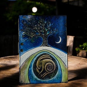 May include: A leather journal with a hand-painted design featuring a tree, crescent moon, and a stylized animal figure. The color palette includes blues, greens, and browns, creating a celestial and natural theme. The journal is closed with snap closures.