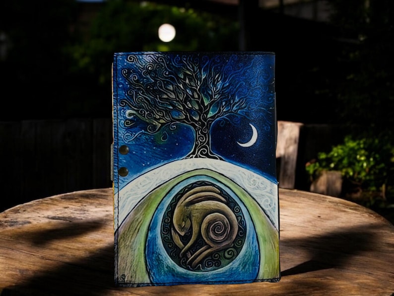 May include: A leather journal with a hand-painted design featuring a tree, crescent moon, and a stylized animal figure. The color palette includes blues, greens, and browns, creating a celestial and natural theme. The journal is closed with snap closures.