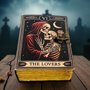 Op de afbeelding: Een ledergebonden dagboek met een tarotkaartontwerp met een skeletpaar dat elkaar omhelst onder een halve maan en sterren. Het dagboek heeft een messing sluiting en goudkleurige pagina's. De kaart luidt "THE LOVERS".