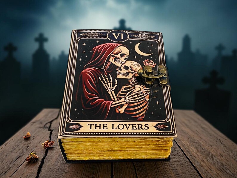 Op de afbeelding: Een ledergebonden dagboek met een tarotkaartontwerp met een skeletpaar dat elkaar omhelst onder een halve maan en sterren. Het dagboek heeft een messing sluiting en goudkleurige pagina's. De kaart luidt "THE LOVERS".