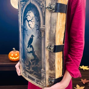 Op de afbeelding: Een groot boek in antieke stijl met een illustratie van een raaf op de omslag. De omslag toont een volle maan, kale bomen en een gotisch boogontwerp. Het boek is gebonden met zwarte leren banden en metalen sluitingen. De pagina's hebben een vintage uitstraling.