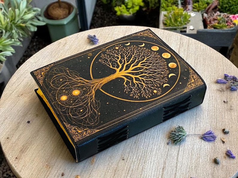 May include: Black journal with a gold tree of life design on the cover. The tree is surrounded by a gold circle with moon phases. The journal has a gold border and gold-edged pages. The journal is on a wooden surface.