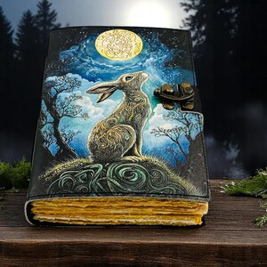 May include: A leather-bound journal with a detailed illustration of a rabbit gazing at a full moon. The cover features a night sky scene with trees and clouds. The journal has a metal clasp and aged-looking pages.
