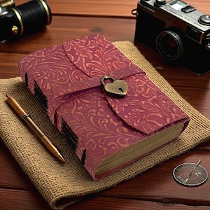 May include: A burgundy leather journal with an embossed floral pattern and a heart-shaped lock. The journal is closed with a strap and sits on a burlap surface, next to a pen and camera.