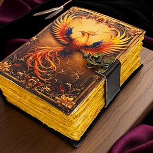 May include: A decorative journal with a phoenix illustration on the cover. The cover features a detailed design with a brass clasp and a black leather strap. The pages have a golden hue, and the journal is resting on a wooden surface.