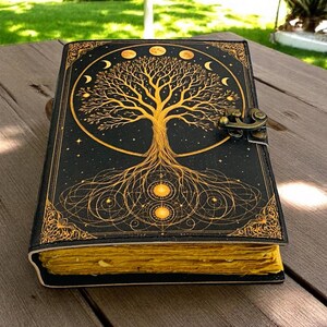 May include: A black journal with a gold tree of life design, moon phases, and stars. The journal has a decorative border and a clasp closure. The pages are yellowed, giving it a vintage look. The journal is on a wooden surface.