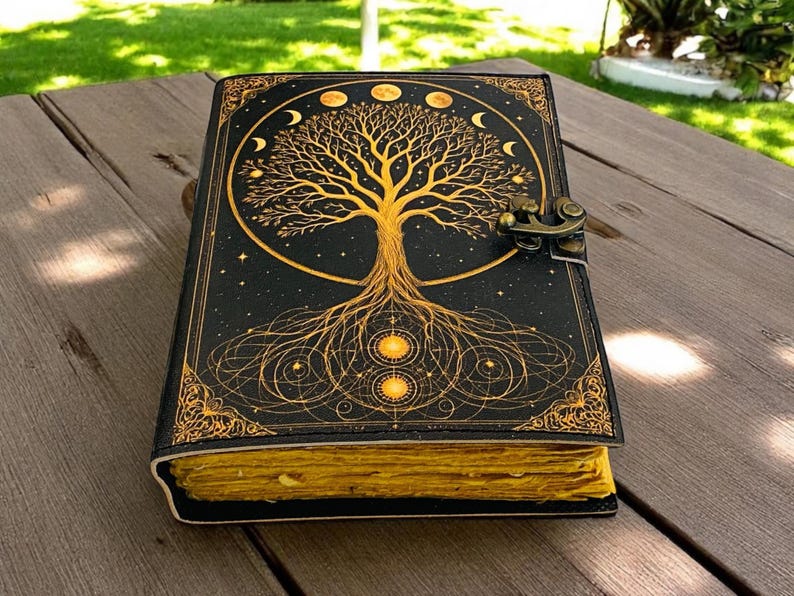 May include: A black journal with a gold tree of life design, moon phases, and stars. The journal has a decorative border and a clasp closure. The pages are yellowed, giving it a vintage look. The journal is on a wooden surface.