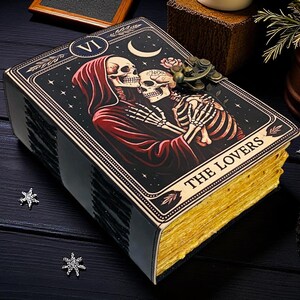 Op de afbeelding: Een tarotkaartdoos met de afbeelding van de kaart "The Lovers". De doos heeft een zwarte achtergrond en goud vergulde randen. De afbeelding toont twee omarmende skeletten, met een rode figuur met capuchon. De tekst "THE LOVERS" staat onderaan.