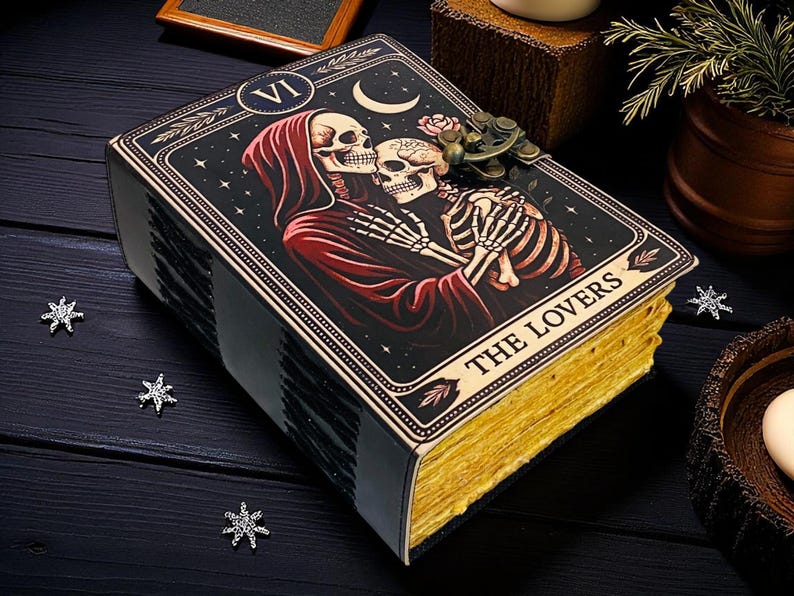 Op de afbeelding: Een tarotkaartdoos met de afbeelding van de kaart "The Lovers". De doos heeft een zwarte achtergrond en goud vergulde randen. De afbeelding toont twee omarmende skeletten, met een rode figuur met capuchon. De tekst "THE LOVERS" staat onderaan.
