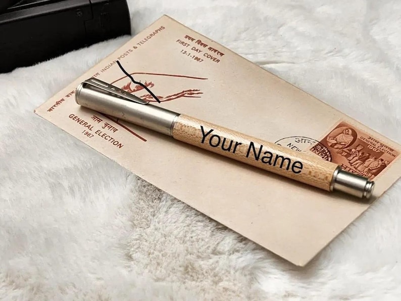K&ouml;nnte beinhalten: Ein personalisierter Holzstift mit dem Text "Your Name" auf dem Schaft. Der Stift hat einen silberfarbenen Clip und eine Spitze. Er liegt auf einem Vintage-Umschlag mit indischen Postmarken und dem Text "GENERAL ELECTION 1967".