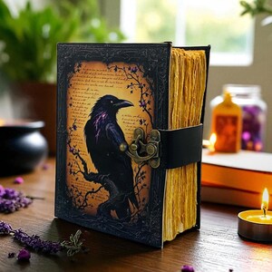 May include: A leather-bound journal with a raven illustration. The cover features a black raven perched on a branch against a parchment-like background. The journal is secured with a black leather strap and a decorative clasp. The pages have a yellowed, aged appearance.