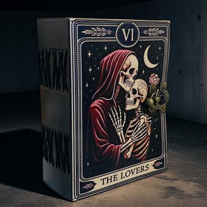 Op de afbeelding: Een zwart, rechthoekig notitieboek met een tarotkaartontwerp. De kaart toont een skeletpaar in een romantische omhelzing tegen een sterrenhemel. De kaart is gelabeld "THE LOVERS" en heeft een metalen sluiting.