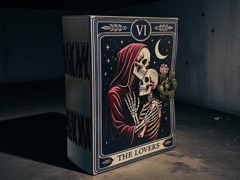 Op de afbeelding: Een zwart, rechthoekig notitieboek met een tarotkaartontwerp. De kaart toont een skeletpaar in een romantische omhelzing tegen een sterrenhemel. De kaart is gelabeld "THE LOVERS" en heeft een metalen sluiting.