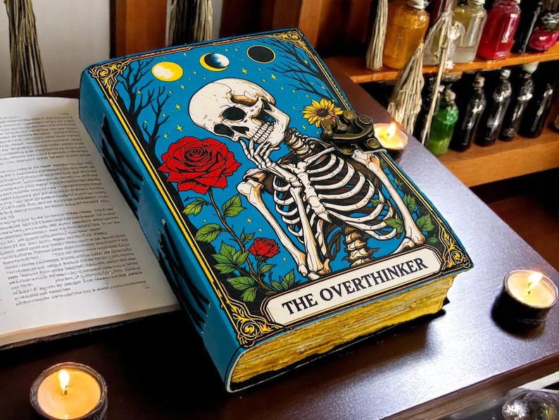 K&ouml;nnte beinhalten: Ein blaues Buch mit goldfarbenen Seiten und einer Skelettillustration. Das Cover zeigt ein Skelett, Rosen und den Text "THE OVERTHINKER". Das Buch liegt offen neben einer brennenden Kerze.