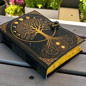 May include: A black leather journal with a gold tree of life design on the cover. The journal has gold-edged pages and a metal clasp. The tree design includes phases of the moon and stars. The journal is closed and resting on a wooden surface.