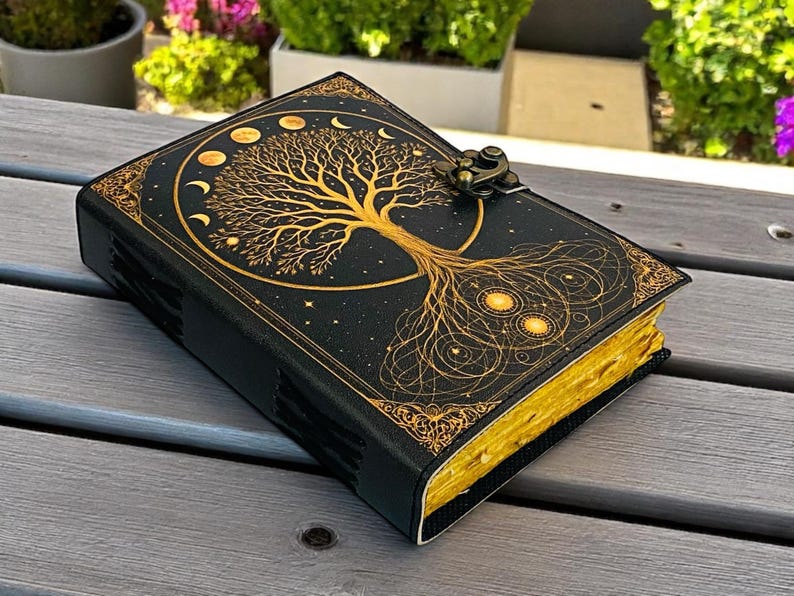May include: A black leather journal with a gold tree of life design on the cover. The journal has gold-edged pages and a metal clasp. The tree design includes phases of the moon and stars. The journal is closed and resting on a wooden surface.