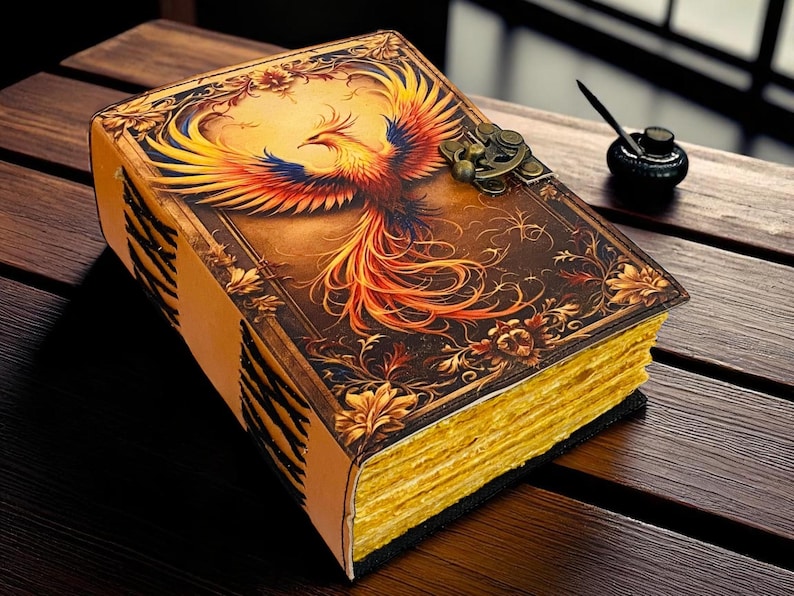 May include: A leather-bound journal with a phoenix illustration. The cover features a detailed image of a colorful phoenix with outstretched wings. The journal has a clasp closure and gold-colored, aged-looking pages. The journal is on a wooden surface.