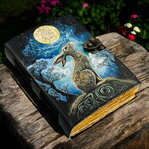 May include: A leather-bound journal with a hand-painted illustration of a rabbit under a full moon. The cover features a night sky scene with trees and a decorative clasp. The pages have a golden hue.