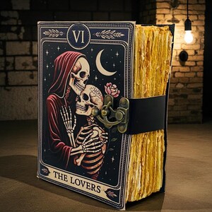 Op de afbeelding: Een tarot-thema dagboek met een zwarte leren riem en sluiting. De omslag toont een skeletpaar in een romantische omhelzing, met een maan en sterren. De pagina's van het dagboek hebben een getextureerde, verouderde uitstraling, met de tekst "THE LOVERS".