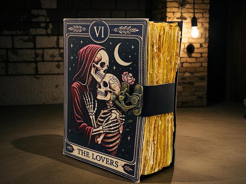 Op de afbeelding: Een tarot-thema dagboek met een zwarte leren riem en sluiting. De omslag toont een skeletpaar in een romantische omhelzing, met een maan en sterren. De pagina's van het dagboek hebben een getextureerde, verouderde uitstraling, met de tekst "THE LOVERS".