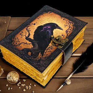 May include: A decorative journal with a raven illustration on the cover, featuring a black leather strap and clasp. The pages have a gold-colored edge. A black feather quill, a small glass bottle, and a decorative ball are also present.