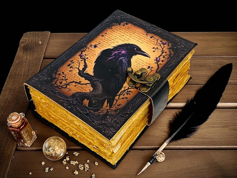 May include: A decorative journal with a raven illustration on the cover, featuring a black leather strap and clasp. The pages have a gold-colored edge. A black feather quill, a small glass bottle, and a decorative ball are also present.