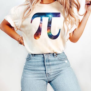 May include: A cream-coloured t-shirt displaying a colourful Pi symbol with a galaxy design. The graphic is a vibrant mix of blues, purples, and oranges. The shirt is paired with light blue denim jeans.