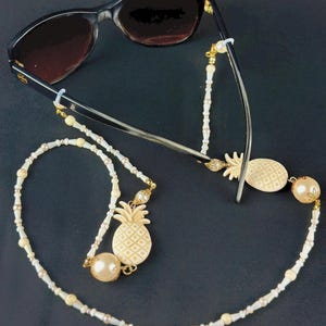 Może przedstawiać: Czarne okulary przeciwsłoneczne z ozdobnym łańcuszkiem do okularów. Łańcuszek zdobią kremowe koraliki, złote akcenty i zawieszki w kształcie ananasa. Do każdego końca łańcuszka przymocowana jest duża kremowa perła.
