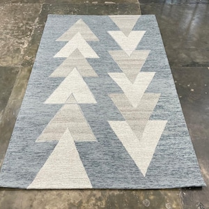 Può includere: Tappeto rettangolare con un motivo geometrico a frecce. Il tappeto presenta uno sfondo azzurro chiaro con file di forme a freccia beige e bianco sporco. Realizzato in materiale tessuto, ideale per il soggiorno.