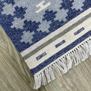 Könnte beinhalten: Nahaufnahme eines gewebten Teppichs mit einem geometrischen Muster in Blau-, Grau- und Weißtönen. Der Teppich hat eine Fransen an einer Kante und eine strukturierte Oberfläche. Das Design umfasst florale und lineare Elemente.