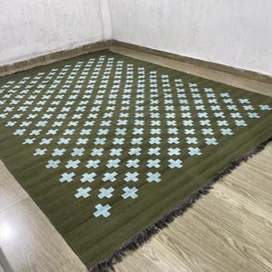 Op de afbeelding: Een rechthoekig olijfgroen tapijt met een herhalend patroon van lichtblauwe kruisen. Het tapijt heeft een gefranjerde rand en ligt op een houten vloer. Dit is een decoratieve vloerbedekking.