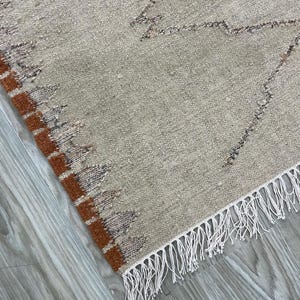 Könnte beinhalten: Beige-brauner gewebter Teppich mit geometrischem Design. Der Teppich hat eine strukturierte Oberfläche, einen braunen Rand und weiße Fransen an einer Kante. Das Muster des Teppichs umfasst Zickzacklinien und dreieckige Formen.