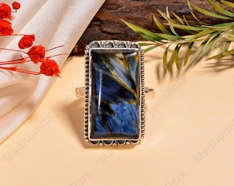 May include: A rectangular silver ring featuring a large, polished blue gemstone with swirling patterns. The gemstone is set in a decorative silver frame with a beaded edge. The ring is displayed on a neutral surface.