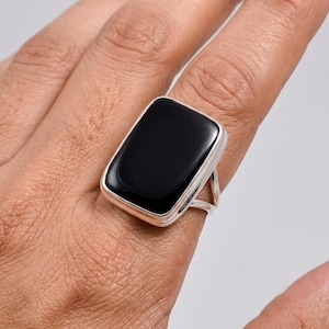 May include: A silver ring featuring a large, smooth, rectangular black stone set in a polished metal bezel. The band is split into two thin, metallic arms that connect to the base of the setting, creating a clean and simple design.