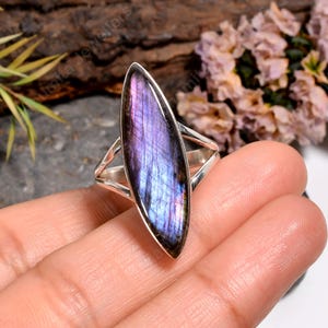 Pode incluir: Anel de prata com uma pedra preciosa de labradorita em forma de marquesa. A pedra apresenta tons iridescentes de roxo, azul e verde. O anel tem um design simples e moderno com haste dividida.