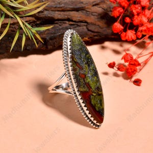 Könnte beinhalten: Ein Silberring mit einem länglichen, oval geformten Stein mit grünen und roten Mustern. Der Stein ist in einer silbernen Fassung mit einem Perlenrand gefasst. Der Ring wird auf einer hellrosa Oberfläche präsentiert.