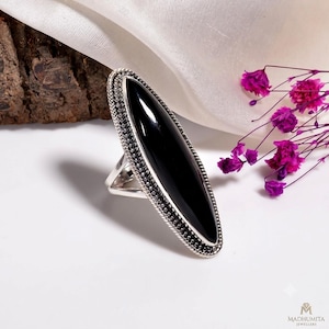 May include: A long, marquise-shaped black onyx gemstone set in a silver ring. The silver band features a detailed beaded and twisted rope texture along the outer edge, framing the smooth, polished black stone in a vertical orientation.