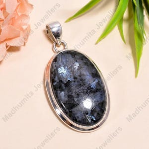 May include: An oval-shaped pendant featuring a polished, dark grey and black speckled gemstone set in a smooth silver frame. The pendant includes a silver bail at the top for attachment to a chain, showcasing a reflective, mottled surface texture.
