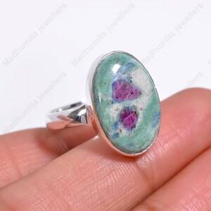 May include: Oval-shaped ring featuring a large, polished gemstone with a light green base, flecks of blue, white, and vibrant red inclusions. The gemstone is set in a silver band. The ring is a piece of jewelry.