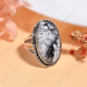 May include: Oval-shaped silver ring featuring a large, polished dendritic opal stone. The stone displays a striking pattern of white and gray, set within a beaded silver frame. The ring's band has a split design.