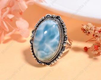 May include: Oval-shaped silver ring featuring a large, light blue gemstone with white marbling. The gemstone is set in a detailed, ornate silver frame. The ring is displayed on a light orange surface, with pink flowers in the background.