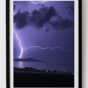 May include: Framed photograph of a dramatic lightning storm over a body of water. The sky is a deep purple, with dark clouds and bright white lightning bolts. The landscape includes a distant island and shoreline.