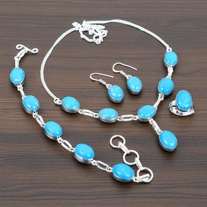 May include: A jewellery set comprising a necklace, bracelet, earrings, and ring. Each item features oval, light blue gemstones set in silver-toned metal. The set is displayed on a dark brown wooden surface.