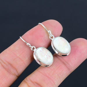 Könnte beinhalten: Ein Paar silberne Ohrhänger mit ovalen, irisierenden weißen Steinen. Die Steine sind in eine silberne Lünette gefasst und haben ein perlmuttartiges Aussehen. Die Ohrringe werden vor einem dunklen Hintergrund gezeigt.
