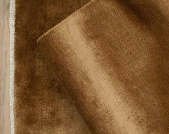 May include: A close-up of a brown rug with a soft, plush texture. The rug is partially rolled up, revealing the underside. The edges have a fringe detail. The rug is a warm, earthy tone, suitable for home decor.