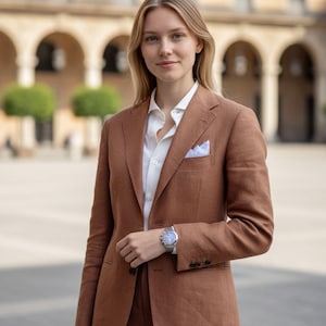 K&ouml;nnte beinhalten: Eine Frau in einem braunen Anzug mit wei&szlig;em Hemd und hellblauem Einstecktuch. Der Anzug hat eine gute Passform. Eine silberne Armbanduhr ist zu sehen. Im Hintergrund ein Geb&auml;ude mit B&ouml;gen.