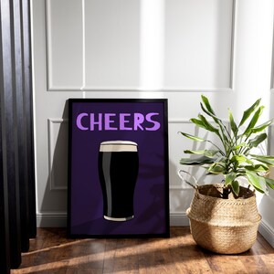 May include: Framed art print featuring a pint of dark beer with a white head, set against a purple background. The word "CHEERS" is written in purple above the beer illustration. A potted plant in a woven basket sits to the right.