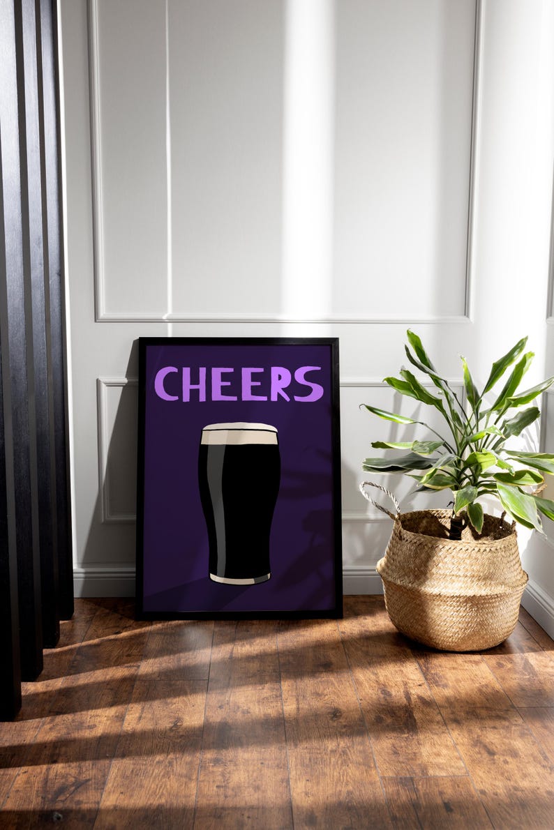 May include: Framed art print featuring a pint of dark beer with a white head, set against a purple background. The word "CHEERS" is written in purple above the beer illustration. A potted plant in a woven basket sits to the right.